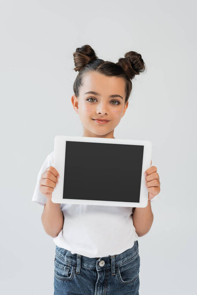 adorable girl with glitter stars on cheeks smiling while holding digital tablet with blank screen isolated on grey