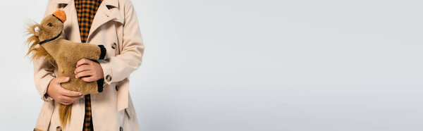 cropped view of girl in stylish trench coat standing with toy horse isolated on grey, banner