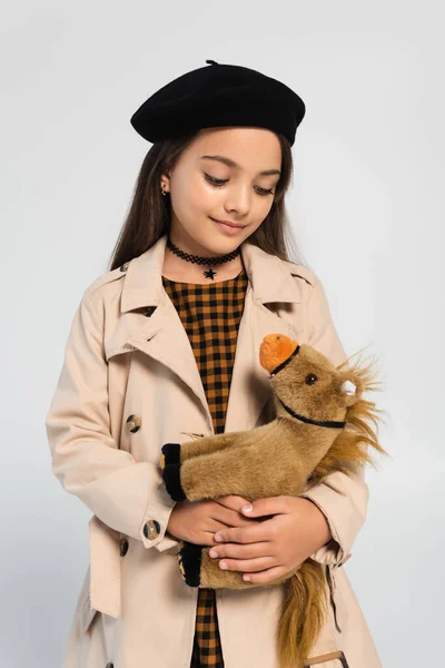 Cheerful Girl Stylish Trench Coat Beret Standing Toy Horse Isolated — Φωτογραφία Αρχείου