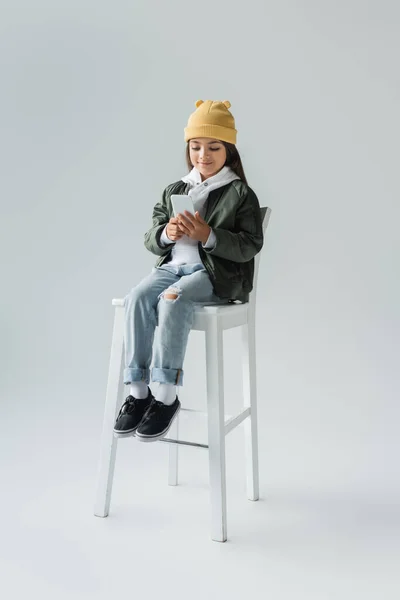 full length of cheerful girl in trendy autumnal outfit sitting on high chair and using smartphone on grey