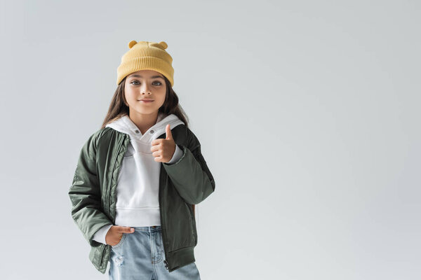 cute girl in beanie hat and stylish autumnal outfit posing with hand in pocket and showing thumb up isolated on grey 