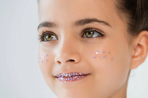 portrait of cheerful girl with sparkling glitter stars on lips and face looking away isolated on white