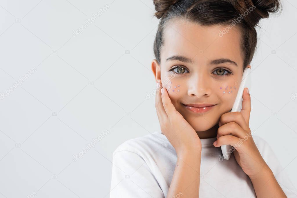 cheerful girl with glitter stars on cheeks talking on smartphone isolated on grey