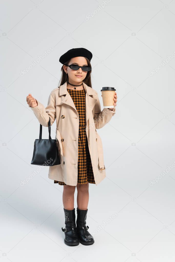 full length of adorable girl in trench coat and sunglasses holding takeaway drink and handbag on grey 