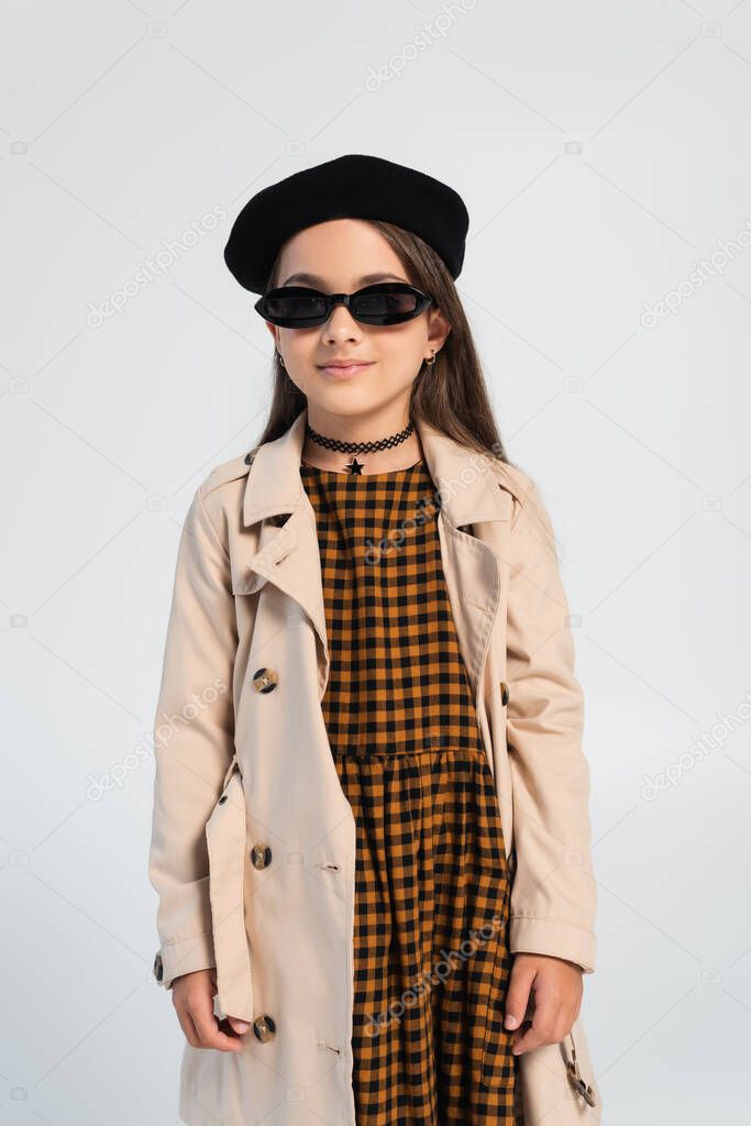 stylish girl in trench coat, beret and sunglasses smiling isolated on grey