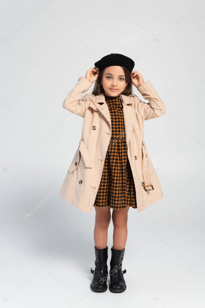 full length of cheerful girl in stylish trench coat adjusting beret while standing on grey