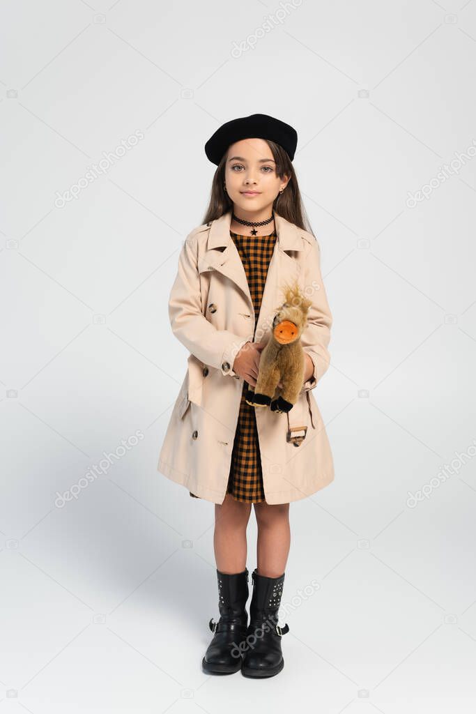 full length of cute girl in stylish trench coat and beret standing with toy horse on grey