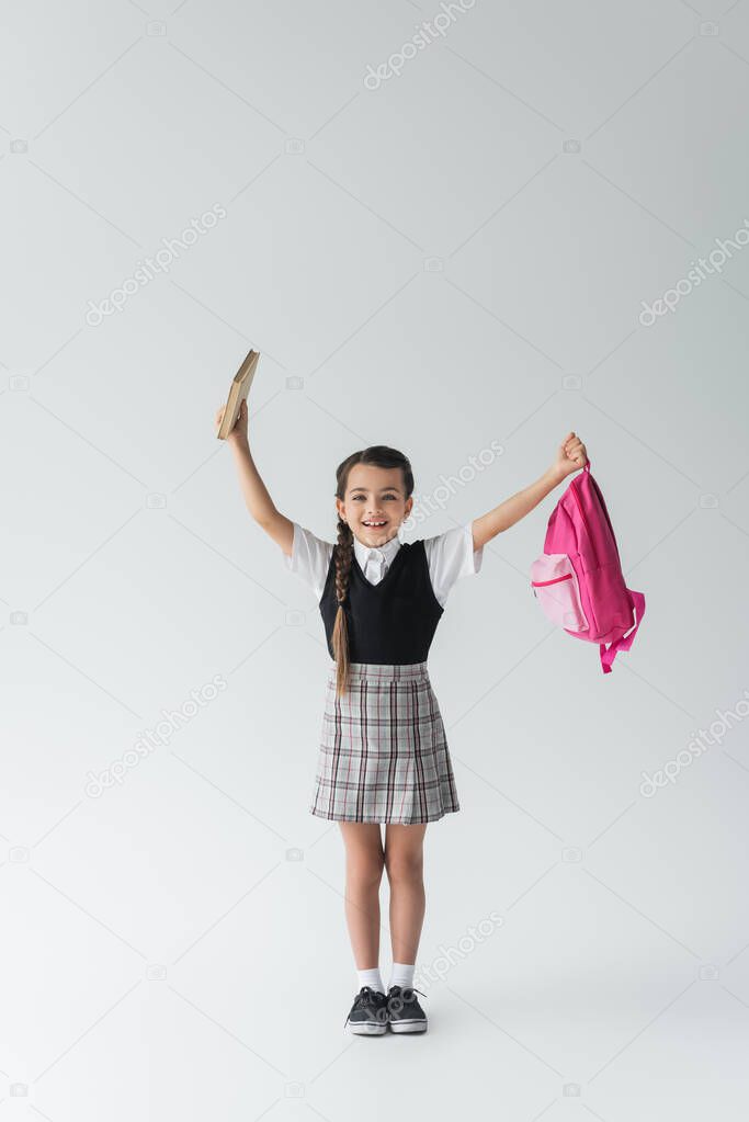 full length of excited schoolgirl in uniform holding pink backpack and book on grey 