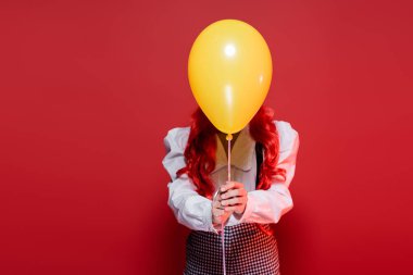 young woman with colored hair obscuring face with yellow balloon on halloween party isolated on red clipart