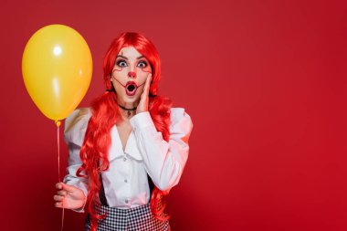 Şaşkın kızıl saçlı palyaço makyajlı kadın elinde sarı bir balon ve kırmızı üzerine dokunmuş bir yüz tutuyor.