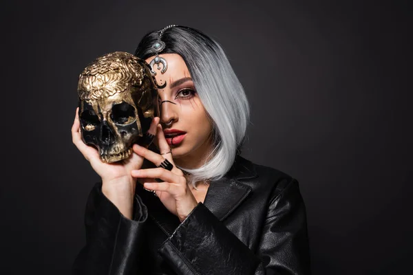 young witch style woman with ash blonde hair obscuring face with spooky golden skull isolated on black