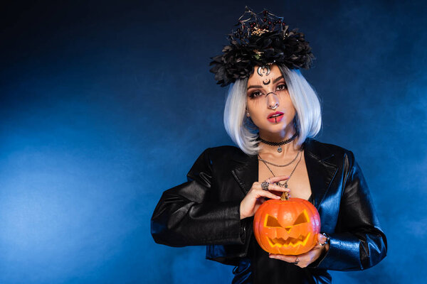 молодая ведьма в черном венке держит резную тыкву рядом с синим туманом на темном фоне