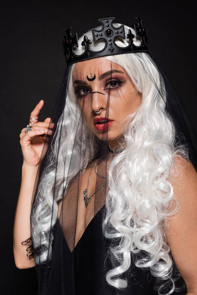 blonde woman in witch halloween costume looking at camera through veil isolated on black