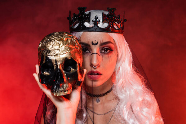 blonde woman with witch makeup wearing black crown and veil while looking at camera near golden skull on red background