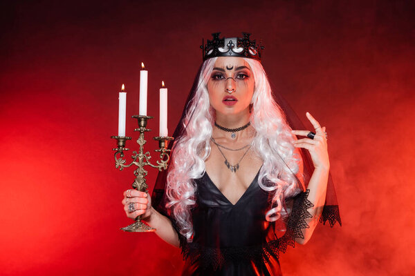 blonde woman in black dress and witch crown holding triple candlestick on background with red smoke