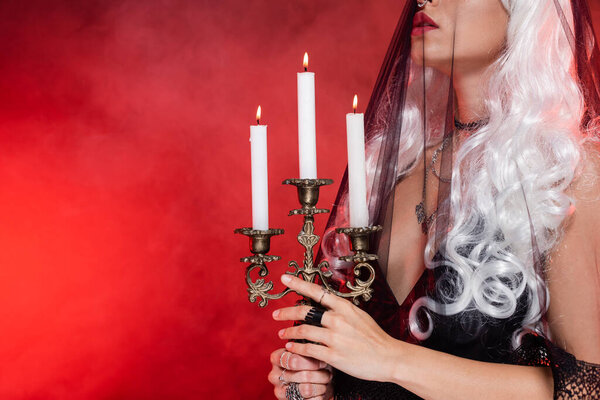 cropped view of blonde witch in black veil holding burning candles on background with red smoke