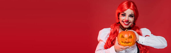 весёлая женщина в стиле клоуна с Джеком Фонарём, смотрящая на камеру, изолированную на красном, баннер