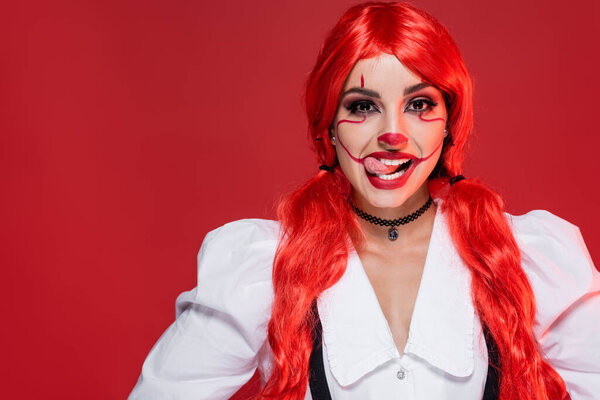 cheerful woman with bright long hair and clown makeup sticking out tongue isolated on red