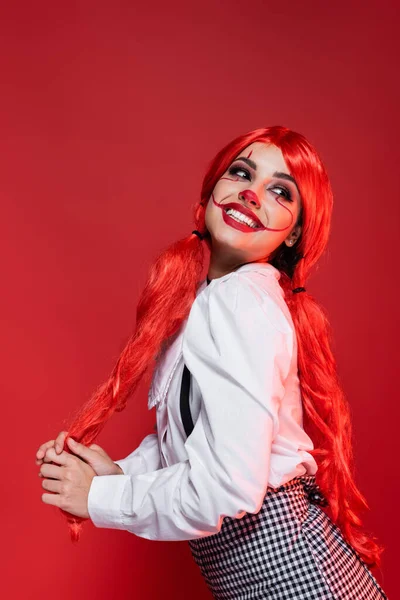 excited woman with colored hair and clown halloween makeup posing isolated on red