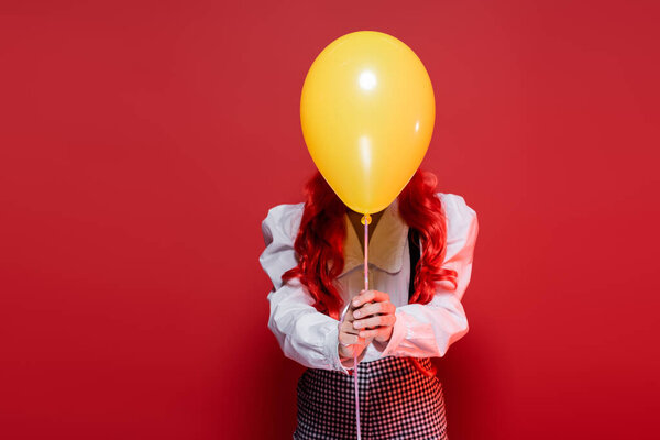 young woman with colored hair obscuring face with yellow balloon on halloween party isolated on red