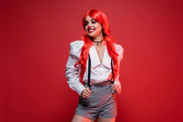 redhead woman with clown makeup posing in white blouse and shorts with suspenders isolated on red