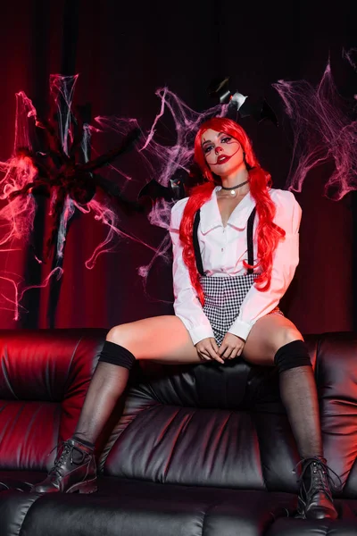seductive redhead woman in clown makeup and black knee socks on leather couch and dark background with spiderweb