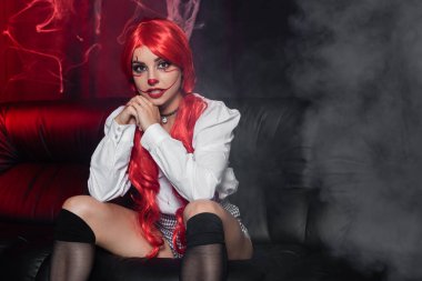 Siyah diz çoraplı ve palyaço makyajlı seksi kızıl kadın deri koltukta oturuyor.