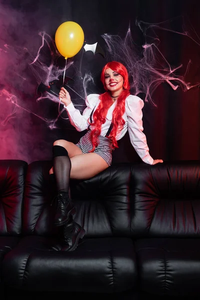 sexy redhead woman in halloween costume holding yellow balloon on leather couch and black background with spiderweb