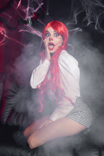 scared woman in halloween makeup touching face while sitting in fog near spiderweb on black background