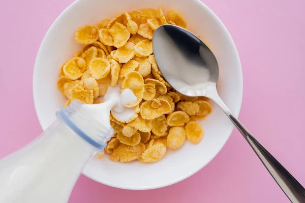 top view of milk pouring from bottle into bowl with corn flakes and spoon on pink