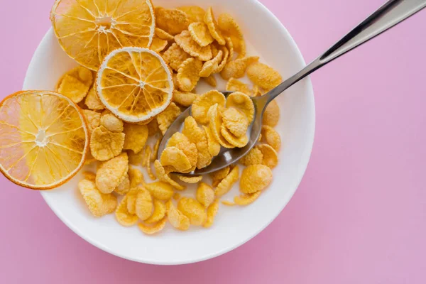 top view of dried oranges in bowl with corn flakes and spoon on pink background