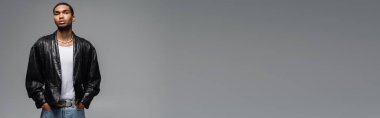 Kotlu ve deri ceketli genç Afrikalı Amerikalı elleri ceplerinde gri pankartta poz veriyor.