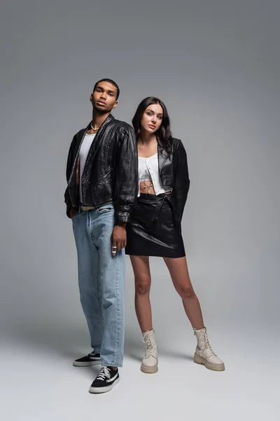 full length of young interracial couple in trendy autumnal outfits standing on grey