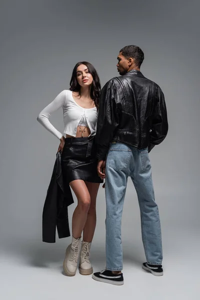 スタイリッシュなアフリカ系アメリカ人男性の近くに黒のジャケットを保持している革のスカートの女性の完全な長さグレー — ストック写真