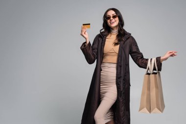Güneş gözlüklü, gülümseyen genç bir kadın alışveriş çantası ve kredi kartı taşıyor.