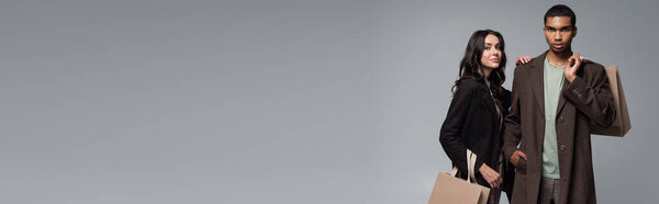 стильная межрасовая пара в осенних нарядах, держащая сумки, изолированные на сером, баннер