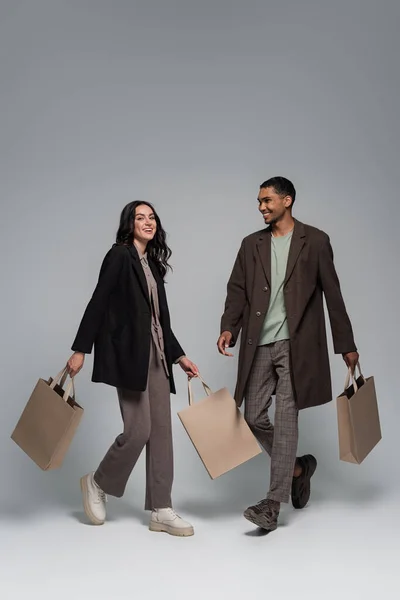全长的快乐的跨种族夫妇 穿着时髦的衣服 提着购物袋 走在灰色的路上 — 图库照片