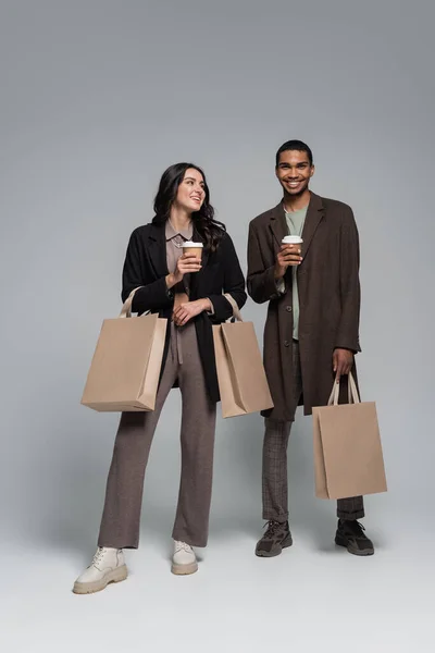 全长的快乐的跨种族夫妇穿着时髦的衣服 手里拿着购物袋和灰色纸杯 — 图库照片
