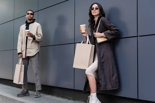 young stylish woman with handbag and paper cup holding shopping bag near african american man