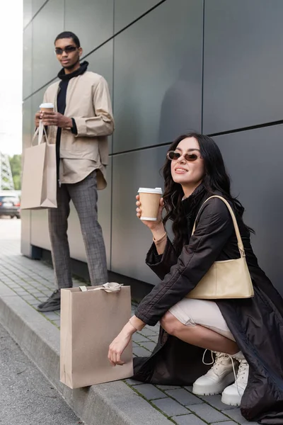 young stylish woman with handbag and paper cup near shopping bag and blurred african american man