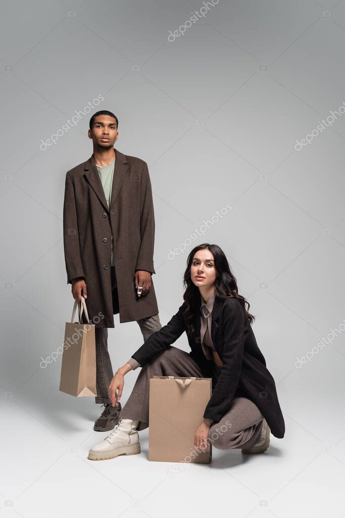 full length of stylish interracial couple in autumnal outfits posing near paper bags on grey