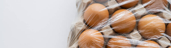 Top view of brown eggs under cellophane in cardboard box on white background, banner 