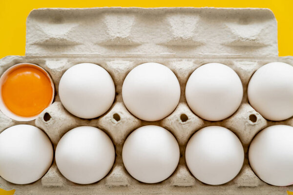 Top view of raw yolk in shell near eggs in carton box on yellow background 