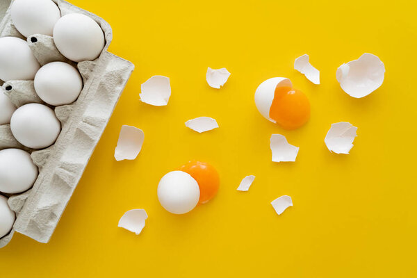 Top view of raw yolks and cracked shells near eggs in tray on yellow background 