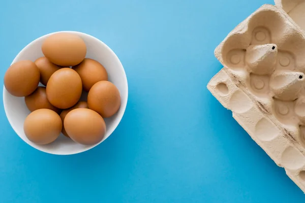 Top view of carton container and chicken eggs in bowl on blue background