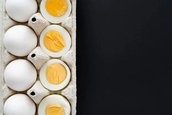 Top view of cut boiled and raw eggs in carton container isolated on black