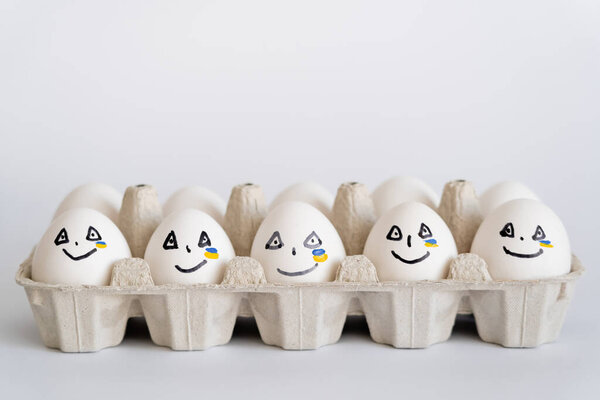 Painted eggs with facial expressions and ukrainian flag in tray on white background 
