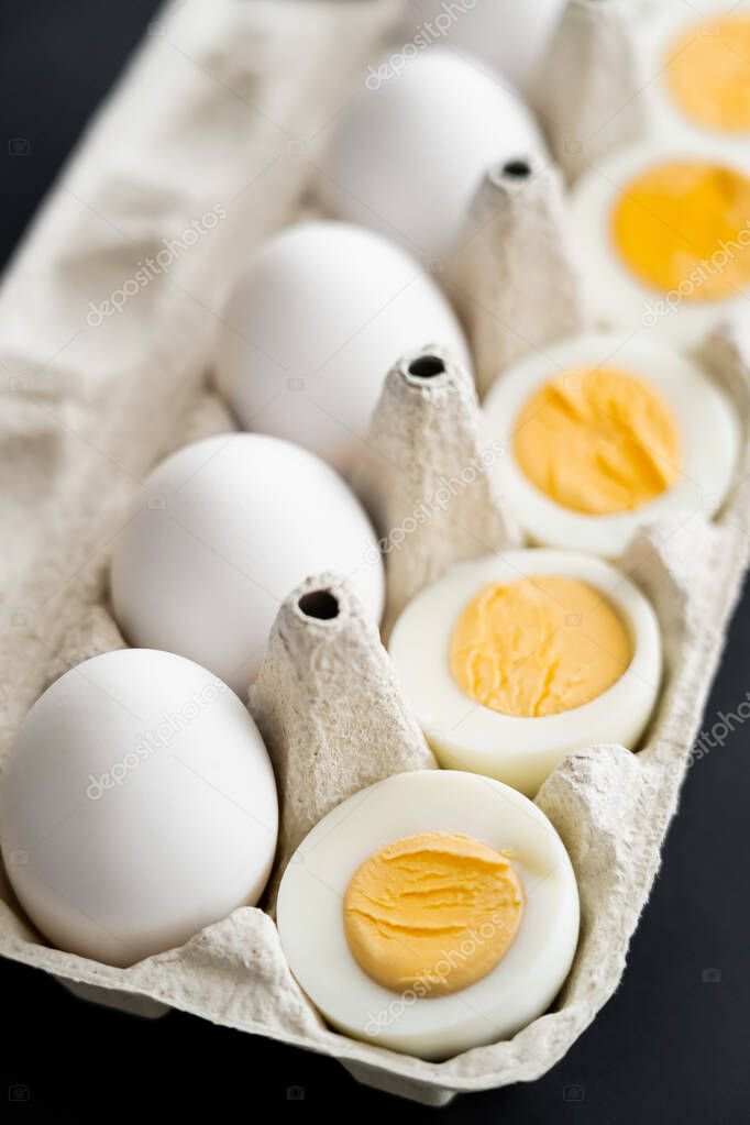 Close up view of cut boiled and raw eggs in blurred carton tray isolated on black 