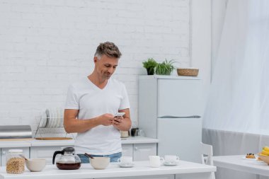 Kahvaltının yanında akıllı telefon ve mutfakta kahve fincanı kullanan gülümseyen adam. 