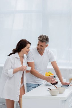 Gülümseyen adam dizüstü bilgisayarını işaret ediyor. Yanında seksi bir eş ve mutfakta kahve. 
